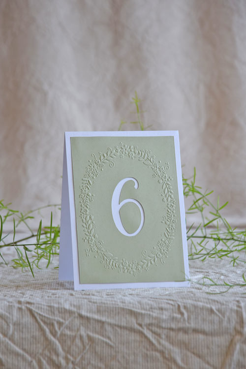 Embossed wedding table number with floral wreath detail, sage panel, and white shimmer border. A romantic folded design for elegant reception tables.